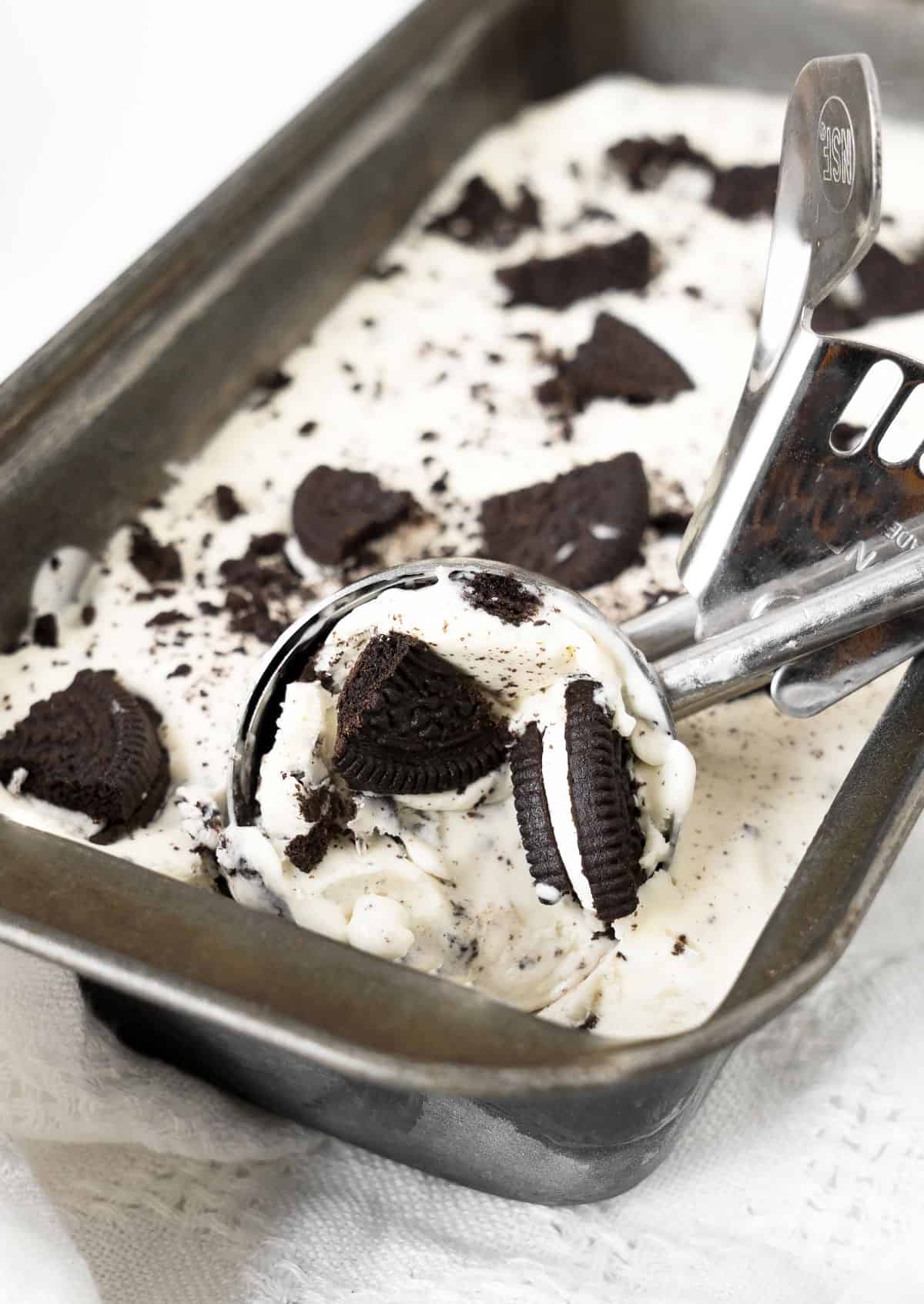 Cookies and cream ice cream in a metal loaf pan with a scoop on a white cloth and background.