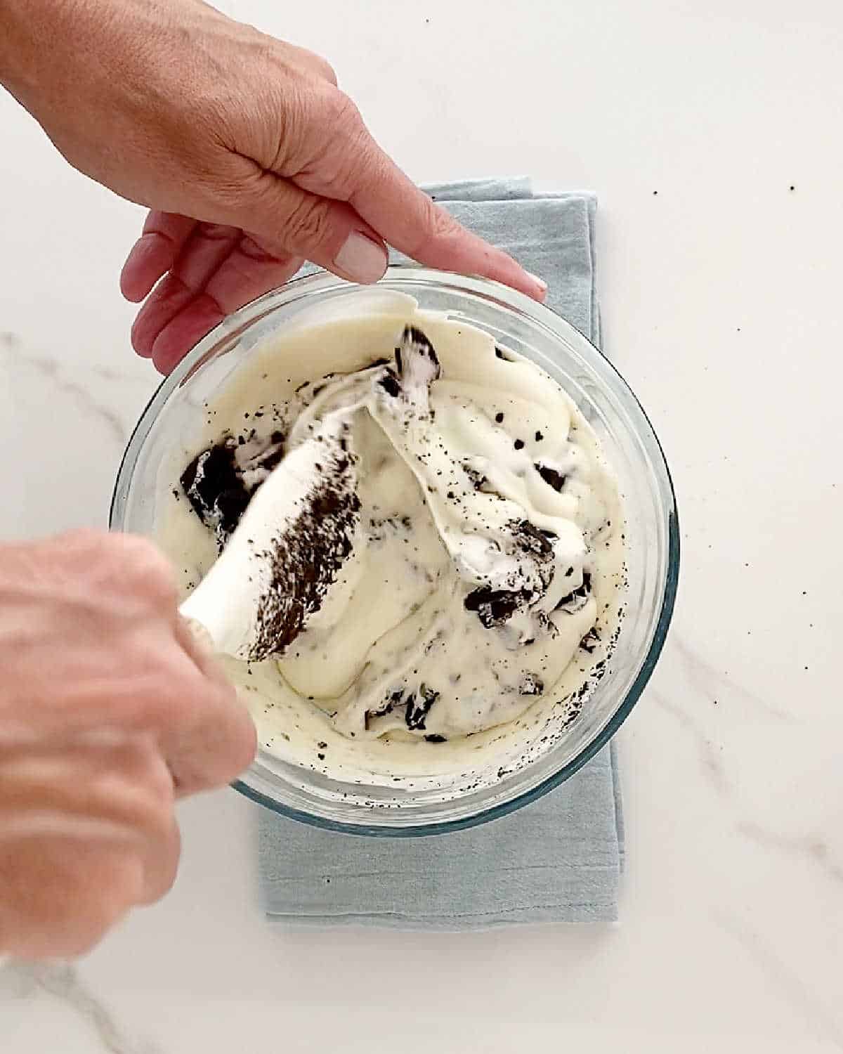 Mixing Oreo cookie ice cream in a glass bowl with a white spatula. White marble surface with light blue cloth.