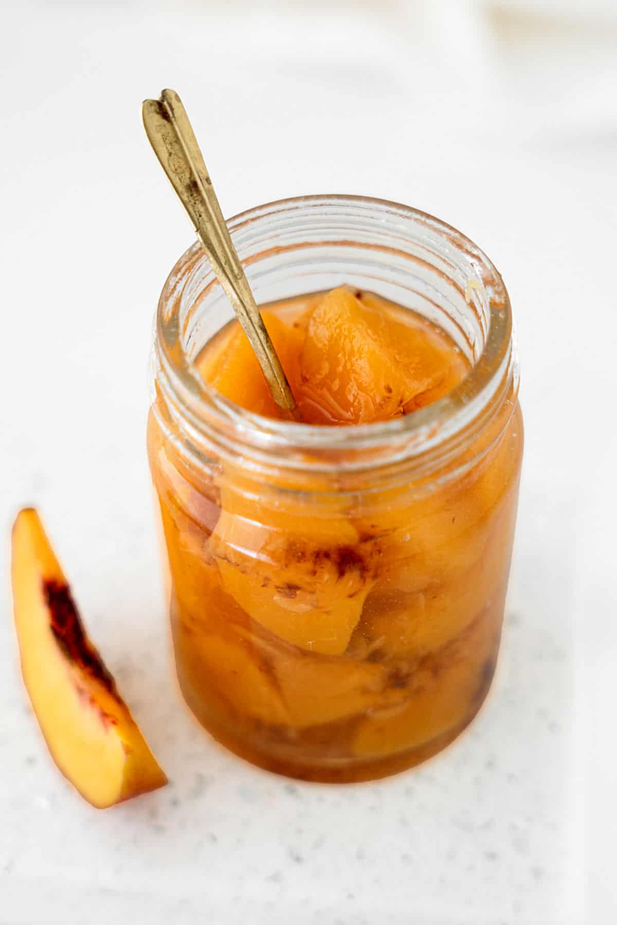 Glass jar with peach compote and a silver spoon on a white surface with a peach wedge.