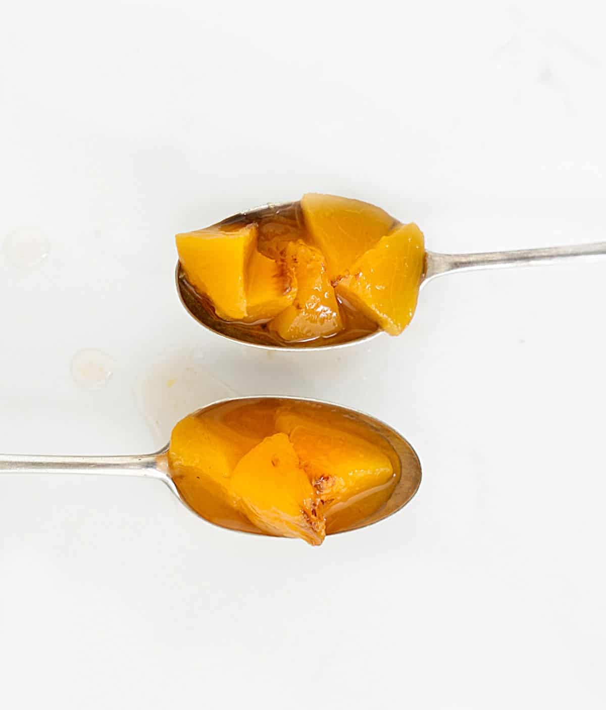 Two vintage silver spoons with peach compote chunks on a white surface.