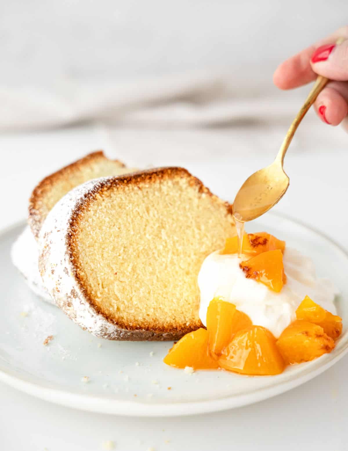 Vanilla cake slices on a white plate with peach compote, cream and syrup drizzling from a gold spoon. White background. 