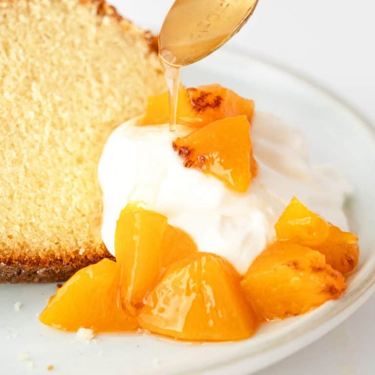 Fresh peach compote with cream on a white plate and syrup drizzling from a gold spoon.