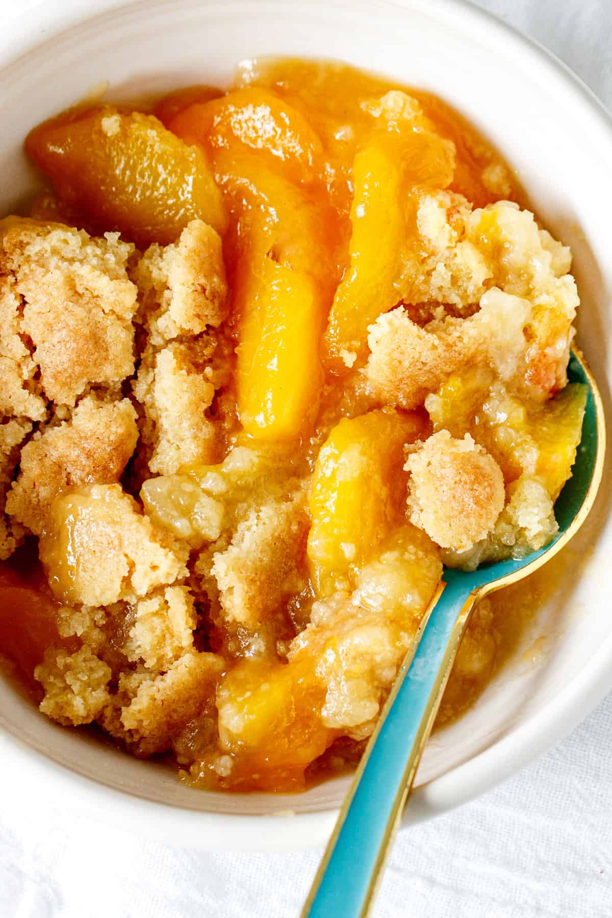 Top view of peach dump cake serving in a white bowl, a teal spoon in it.