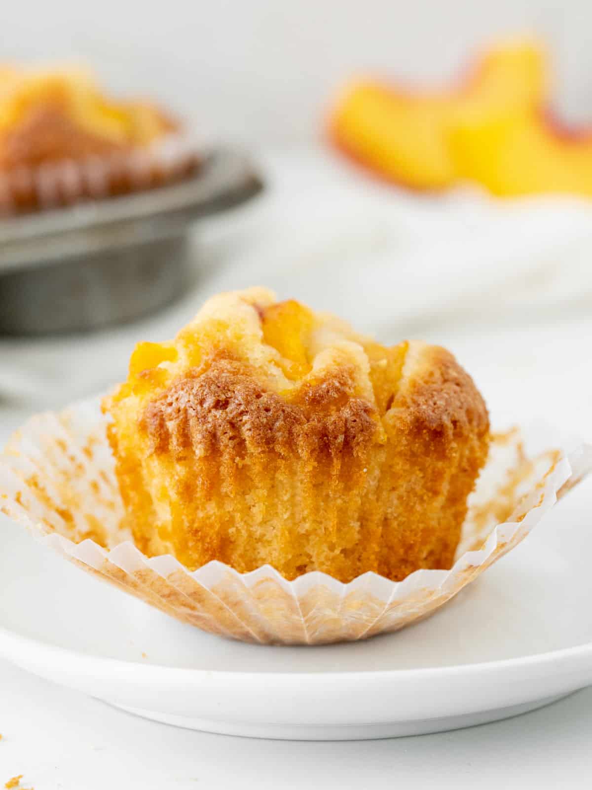 Whole peach muffin in an opened paper liner on a white plate. Muffin pan in the white background. 