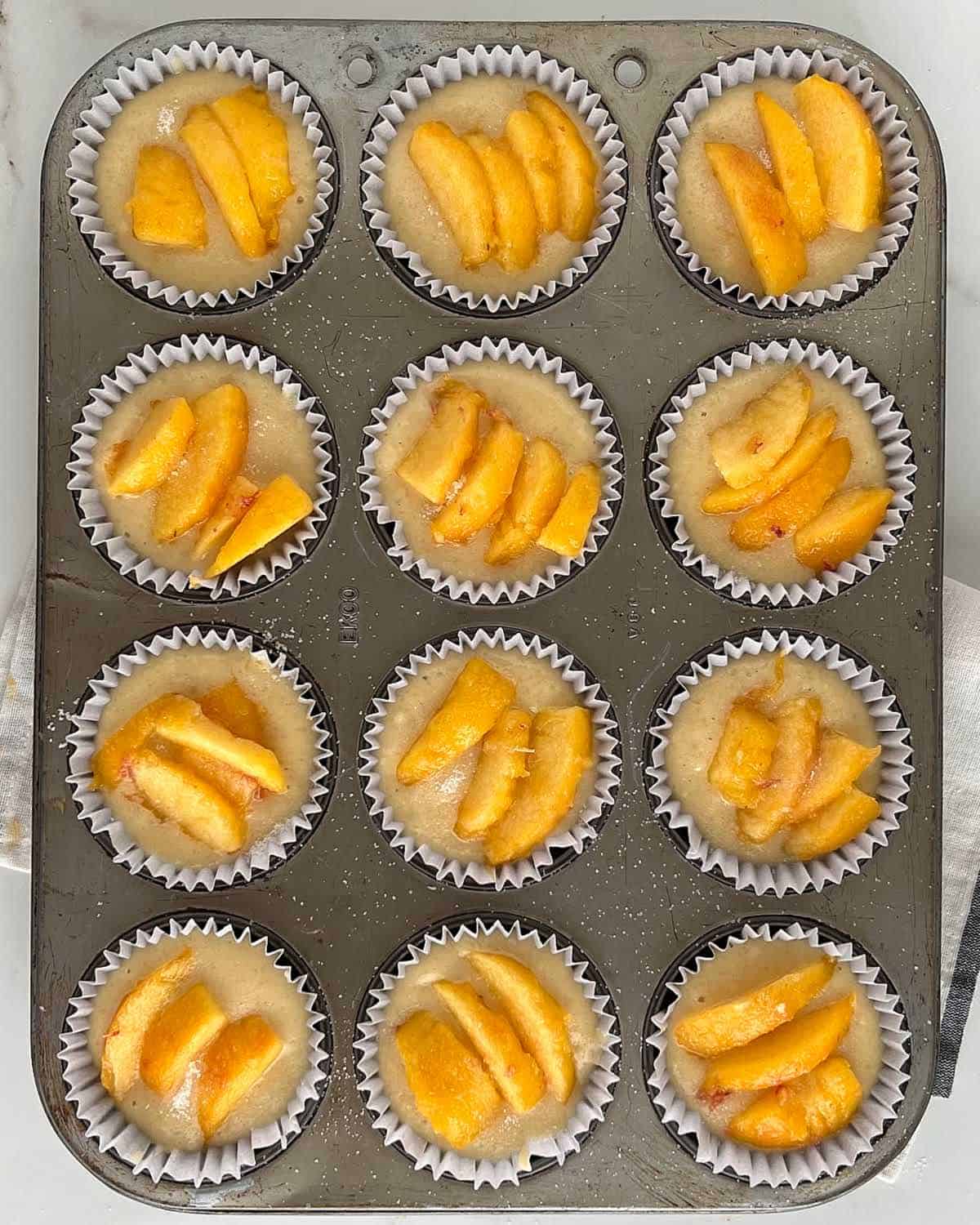 Muffin tin with peach-topped muffin batter in paper liners.