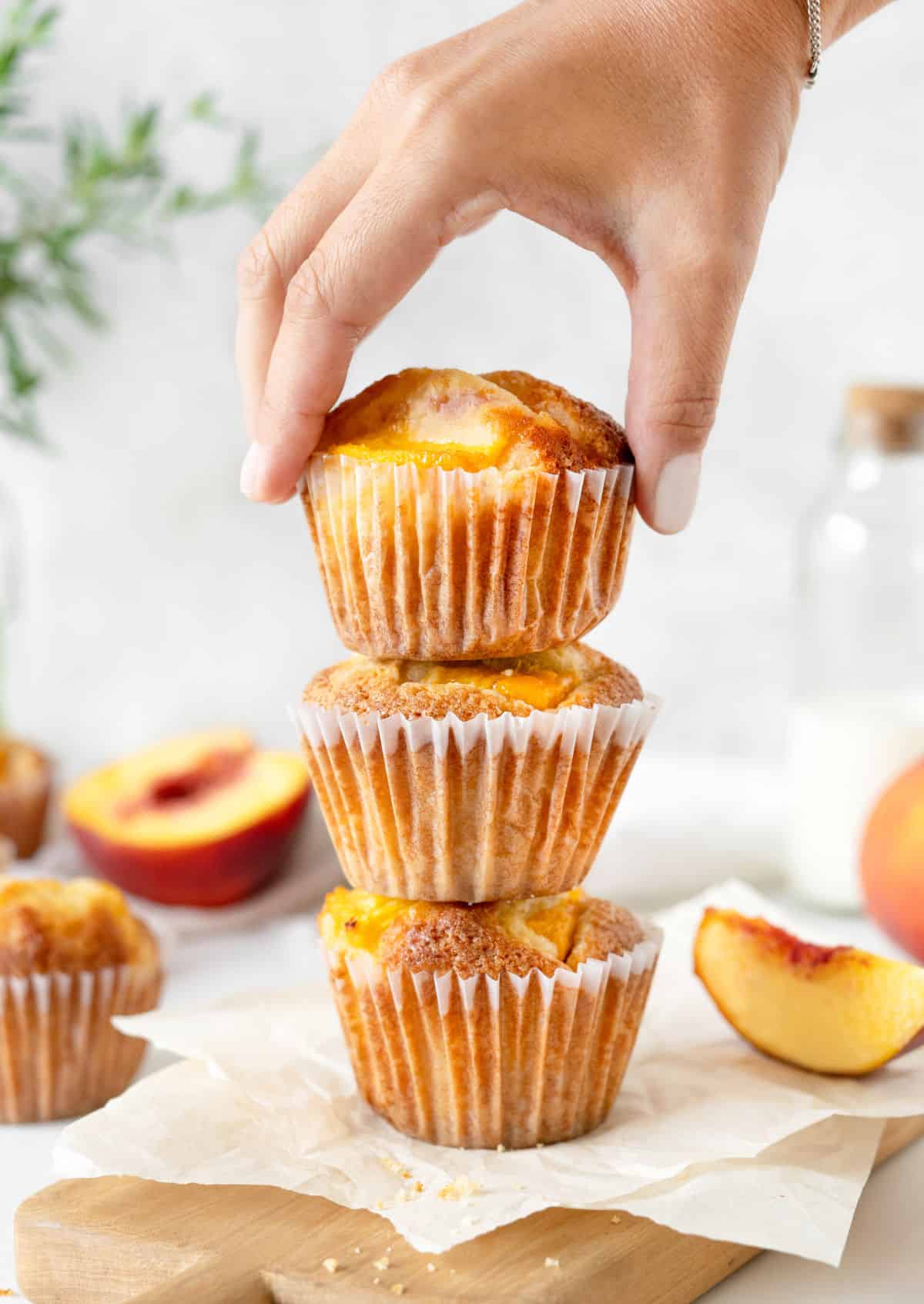 A hand picking top peach muffin from a stack on parchment paper. Fresh peach slices around, grey background.