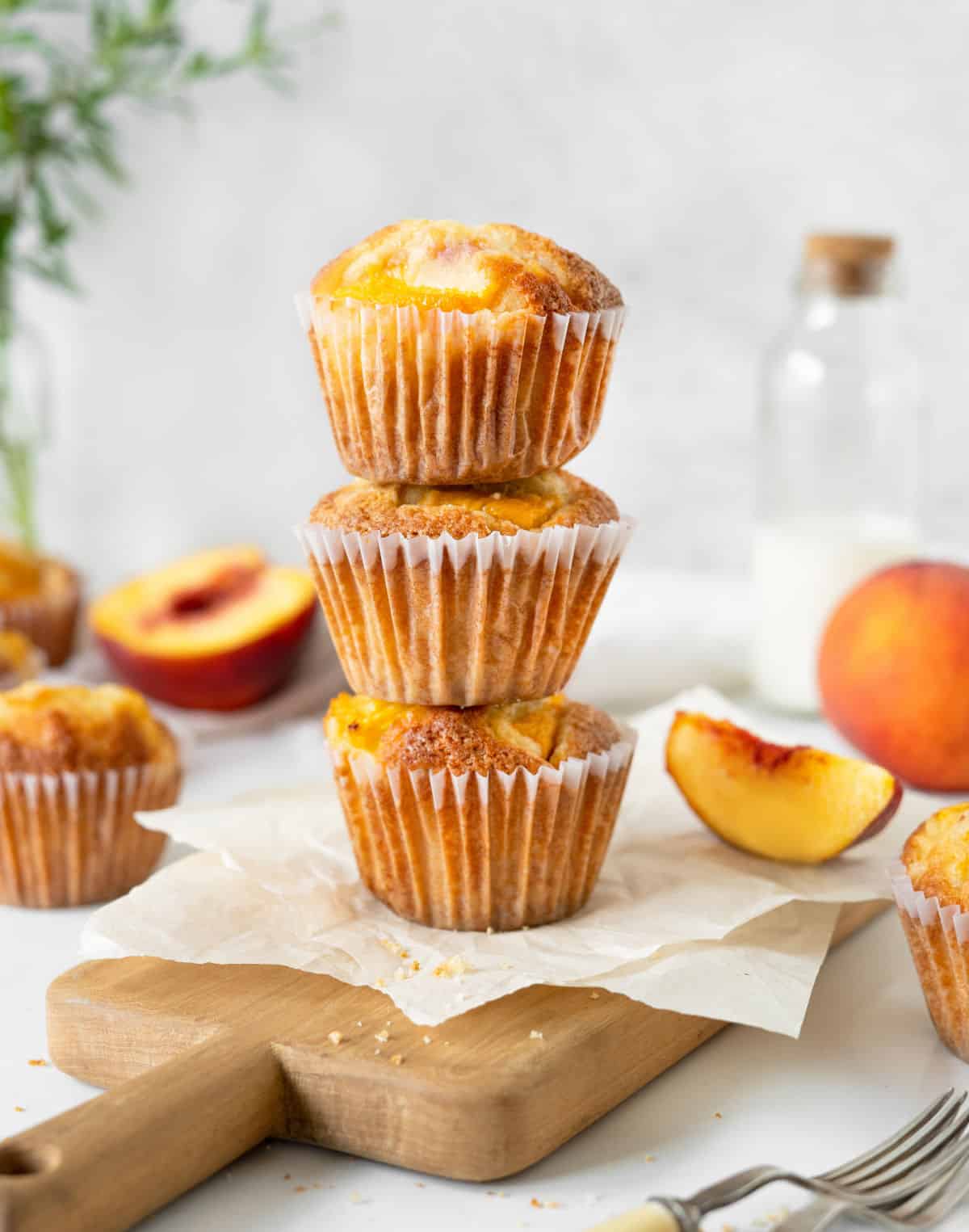 A stack of three peach muffins on beige parchment paper, greyish background, fresh peaches around.