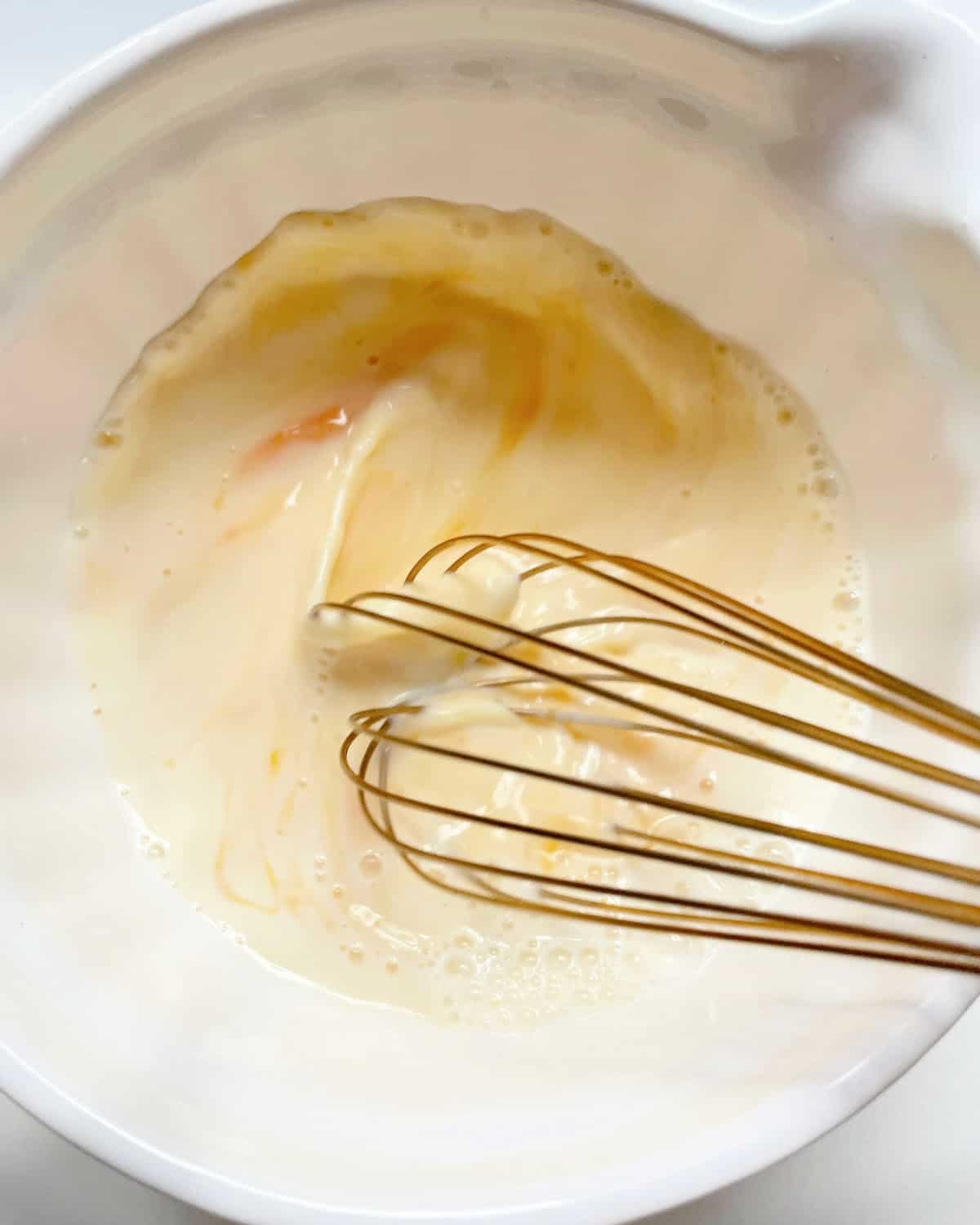 Whisking eggs and milk in a white bowl with a gold-colored whisk. 