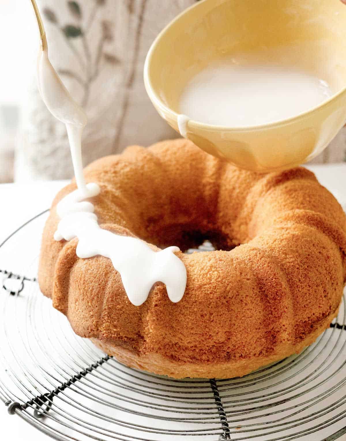 Icing a bundt cake on a wire rack with powdered sugar glaze from a yellow bowl.
