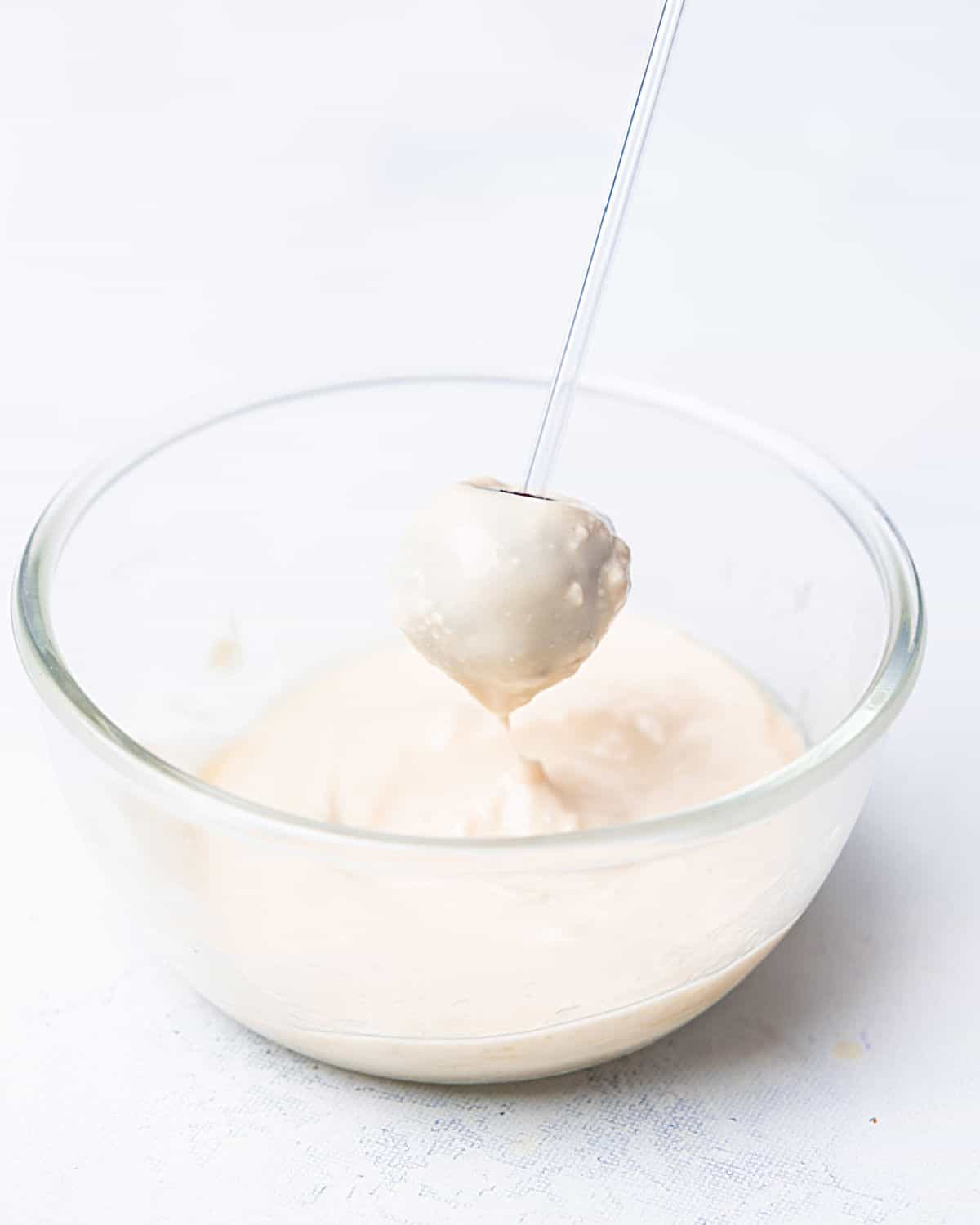 Dipping a cake pop in white chocolate in a glass bowl on a white surface.