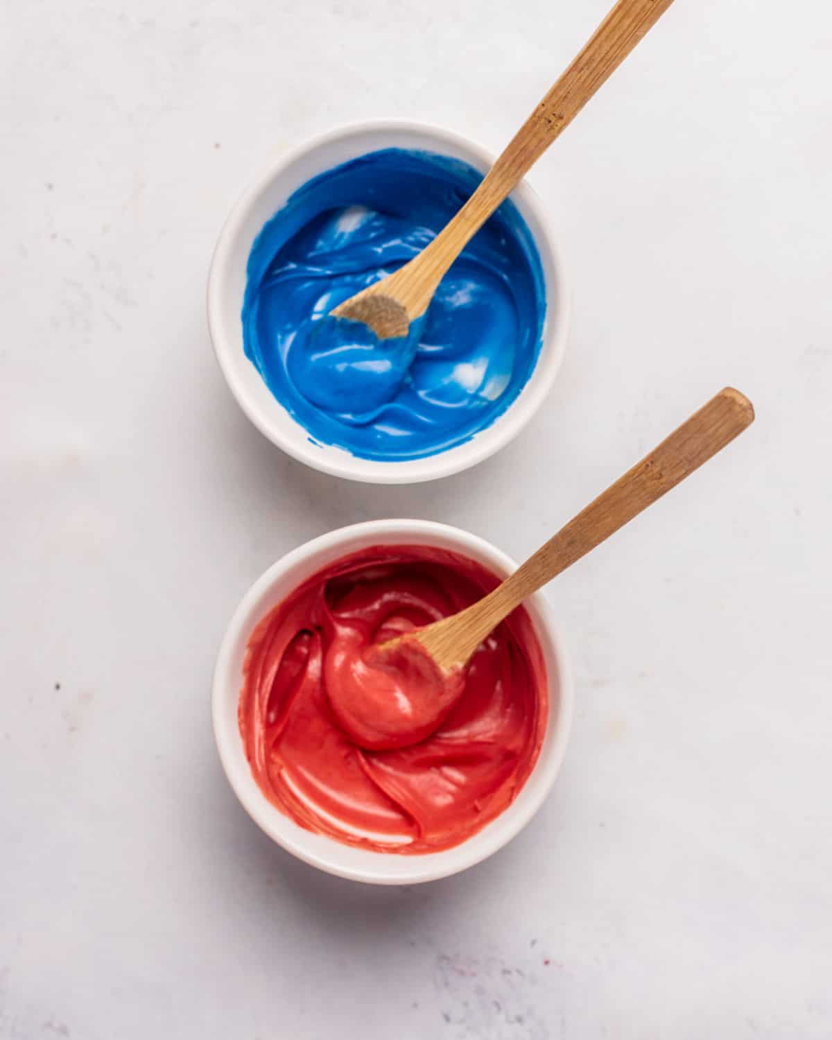 Two white bowls on a grey surface with blue and red colored chocolate and wooden spoons.