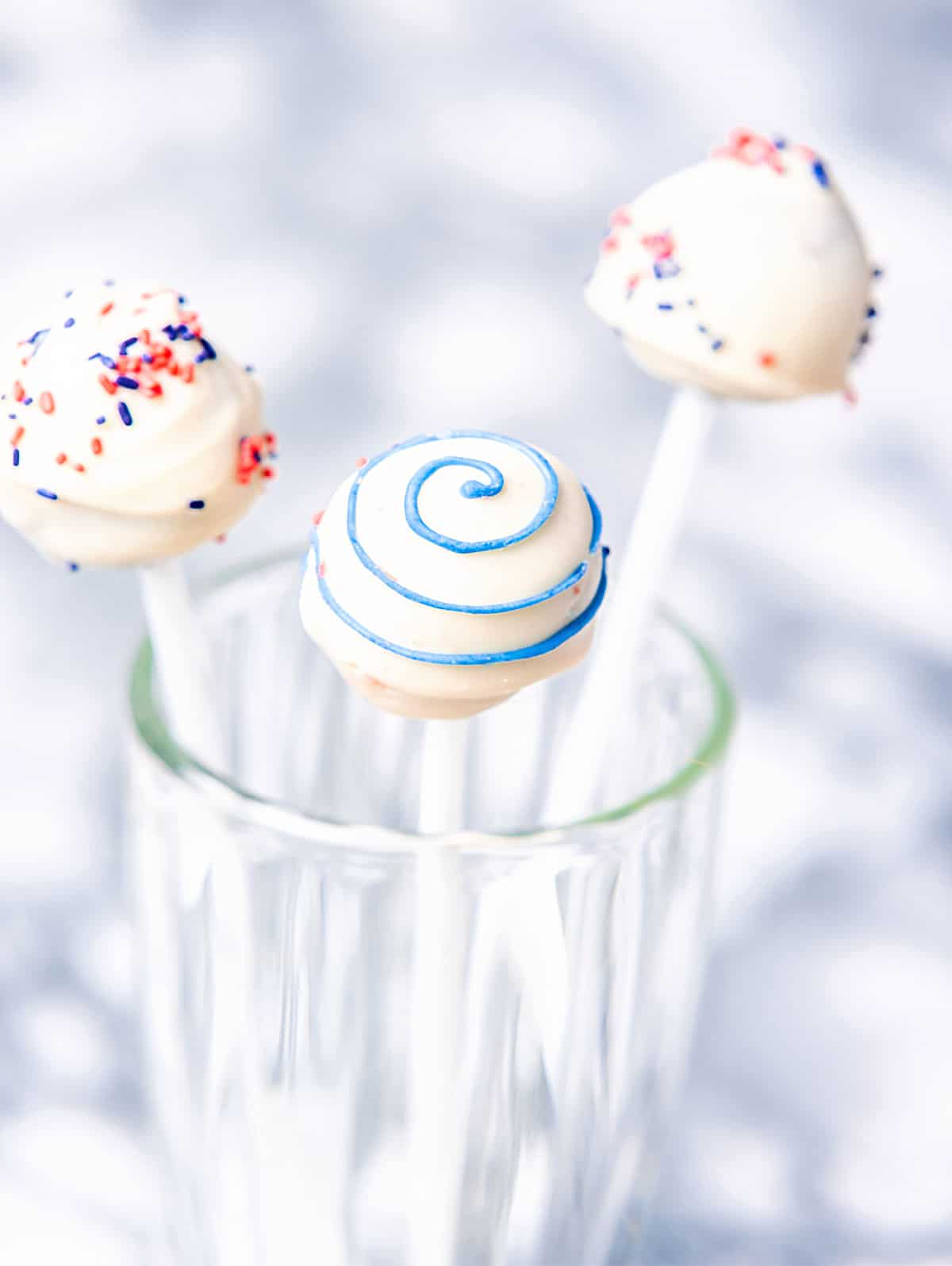 Three white, red, and blue cake pops in a tall glass on a bluish grey background. 