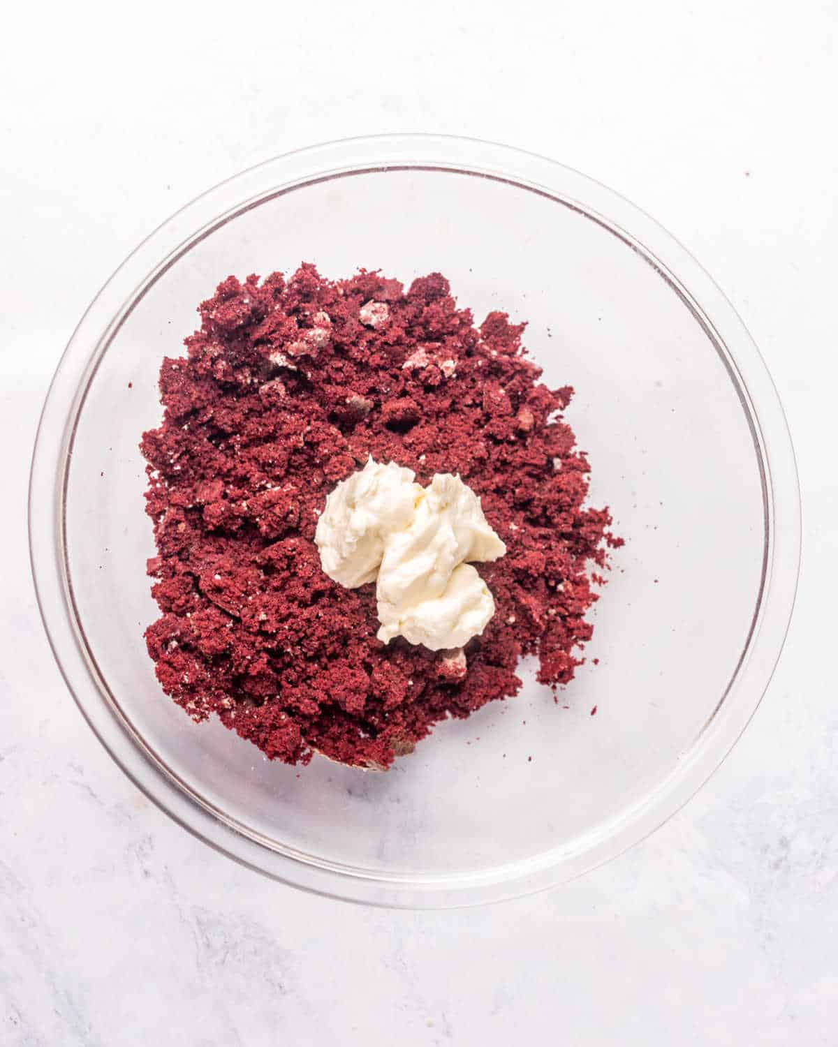 Glass bowl with red velvet cake crumbs and cream cheese dollop on a greyish surface.