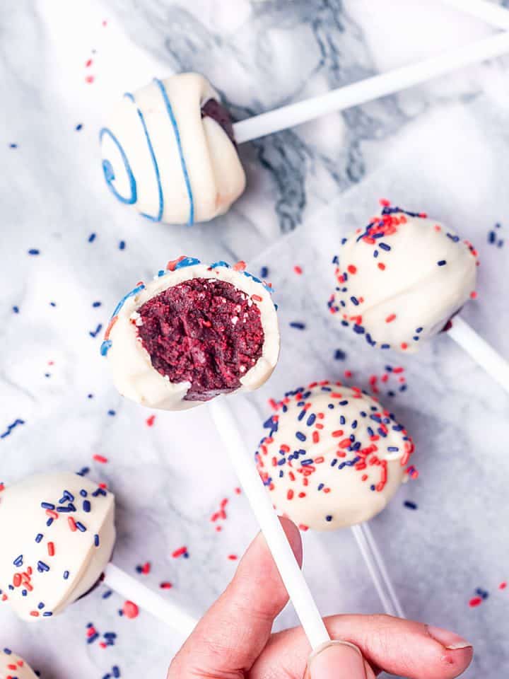 Bluish marble surface with several white chocolate red velvet cake pops with blue and red sprinkles and icing.