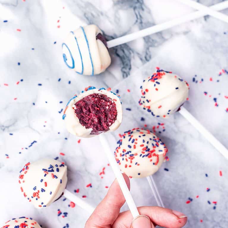 Bluish marble surface with several white chocolate red velvet cake pops with blue and red sprinkles and icing.