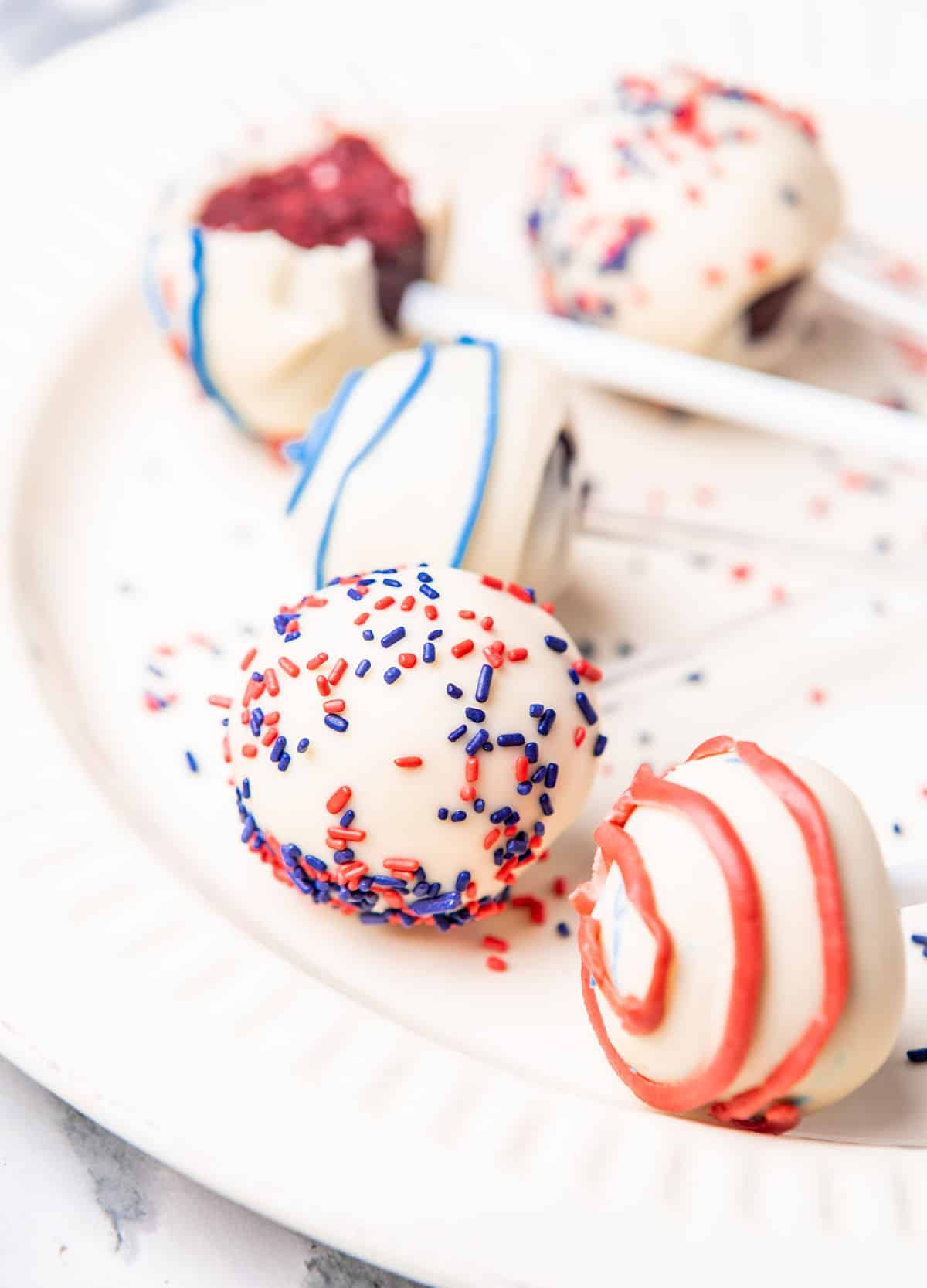 White plate with several white chocolate covered cake pops with red and blue sprinkles and icing.