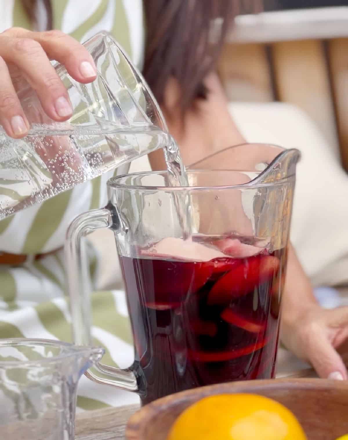 Adding lime soda to a glass with red wine and sliced fruit. Person in green white dress.