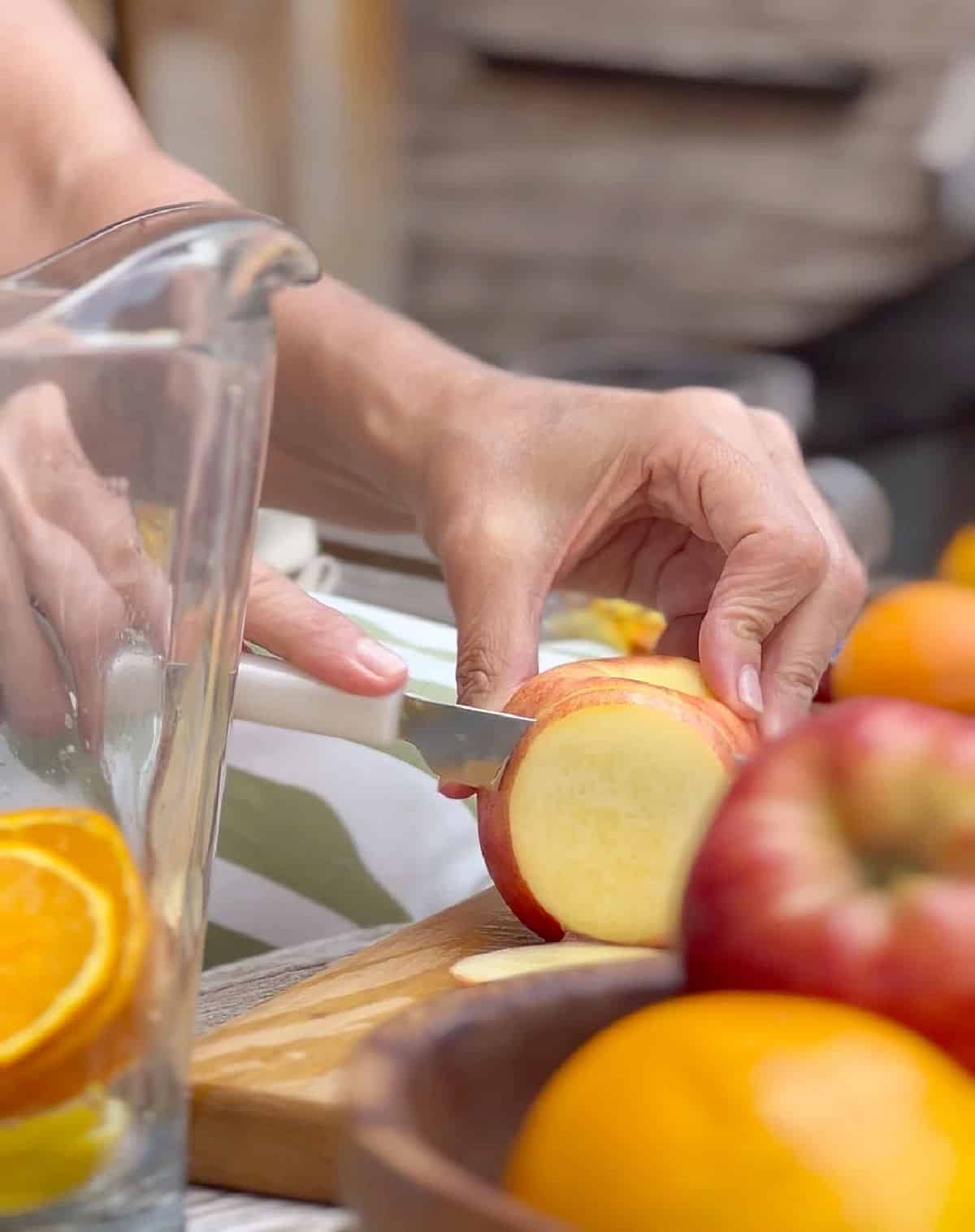Cutting a peach in slices on a board, a pitcher with sliced oranges and bowl of fruit.
