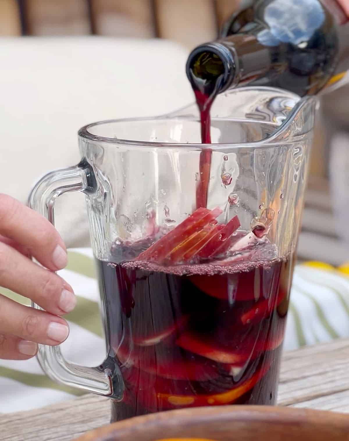 Adding red wine to a glass pitcher with sliced fruit.