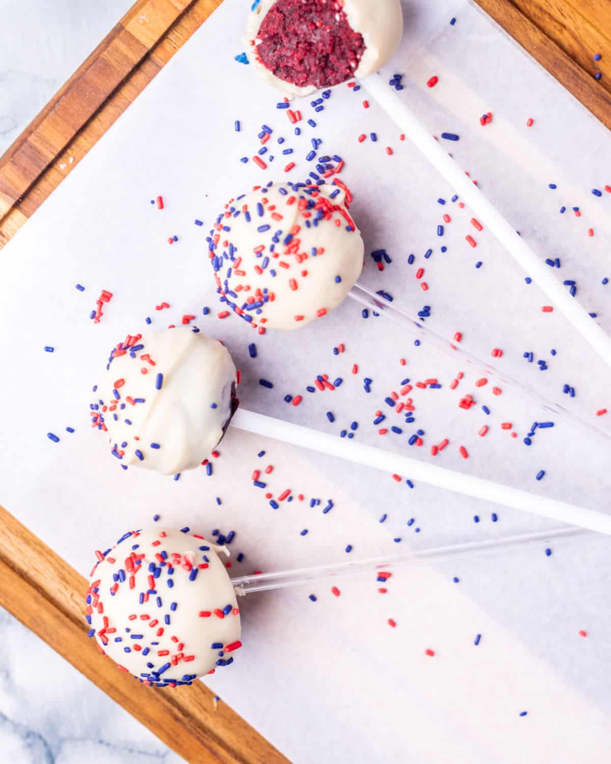 Several white covered cake pops with red and blue sprinkles on parchment paper and a wooden board.