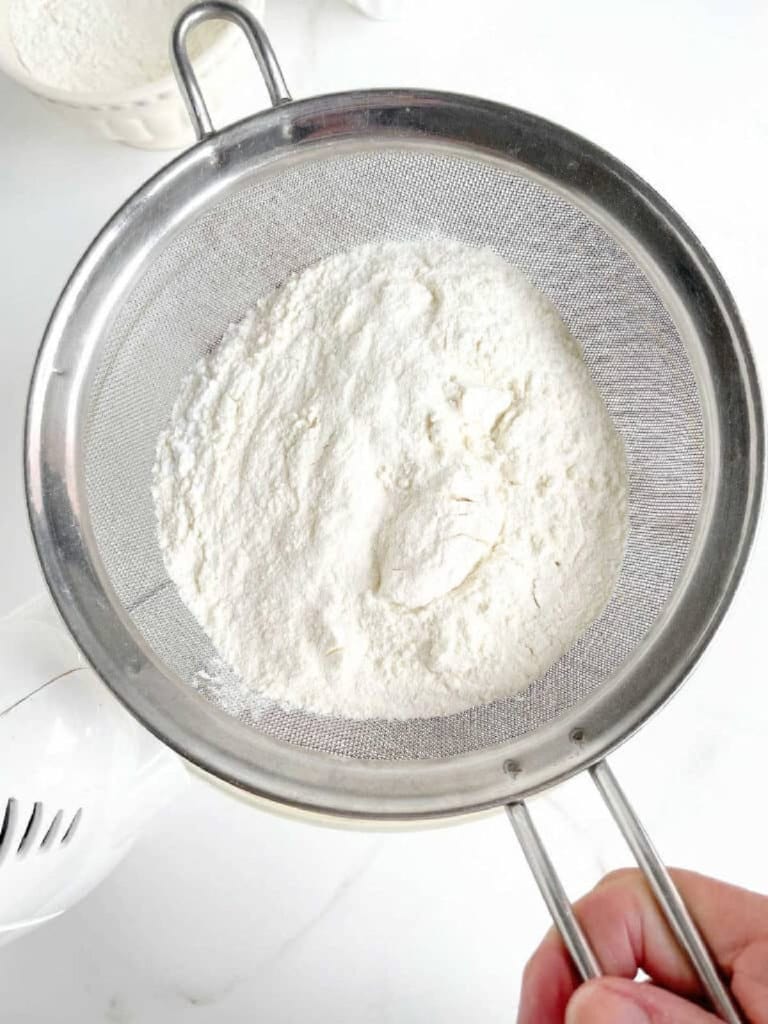 Sifting flour mixture over glass bowl with cake batter. White surface. Top view.