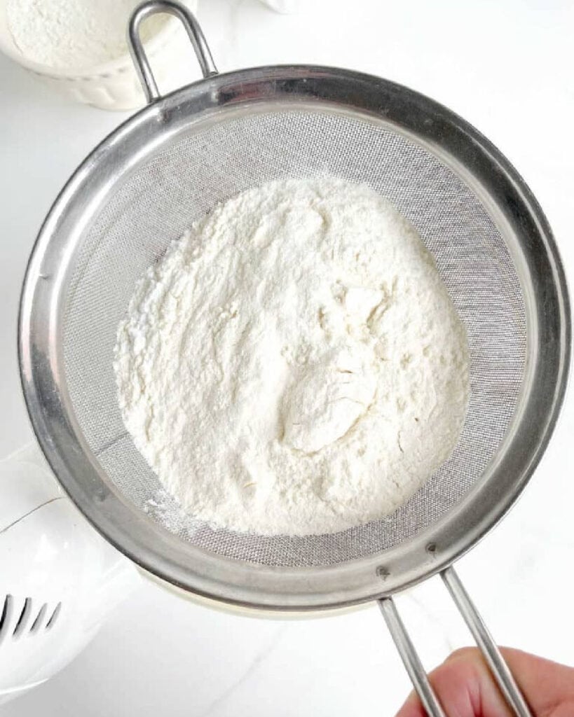 Sifting flour mixture over glass bowl with cake batter. White surface. Top view.
