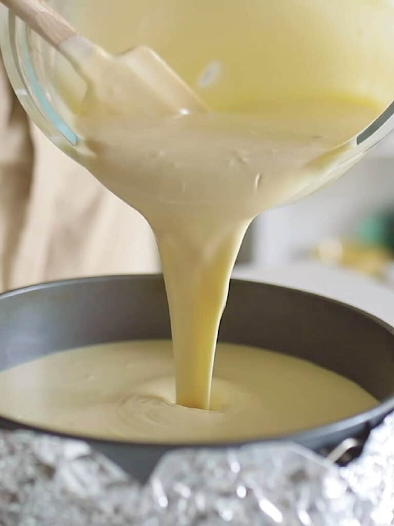 Pouring cheesecake mixture into round cake pan lined with aluminum foil.