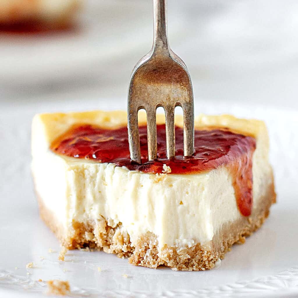 White background and plate with eaten slice of jam covered cheesecake. Silver fork.