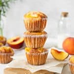Three peach muffins in a stack on parchment paper with greyish background. Fresh peaches around.