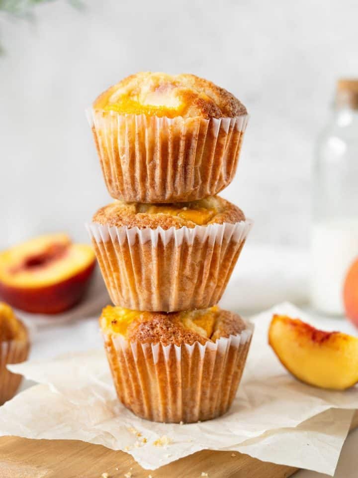 Three peach muffins in a stack on parchment paper with greyish background. Fresh peaches around.