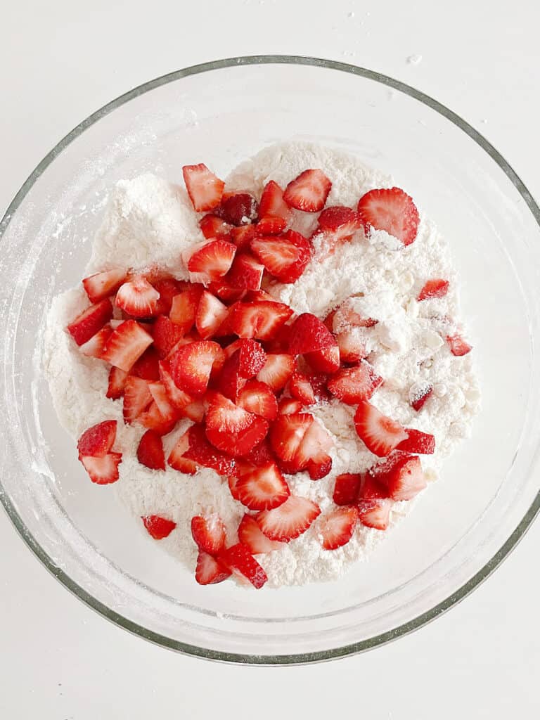 Small strawberry pieces scatter over scone mixture. Glass bowl. White surface.