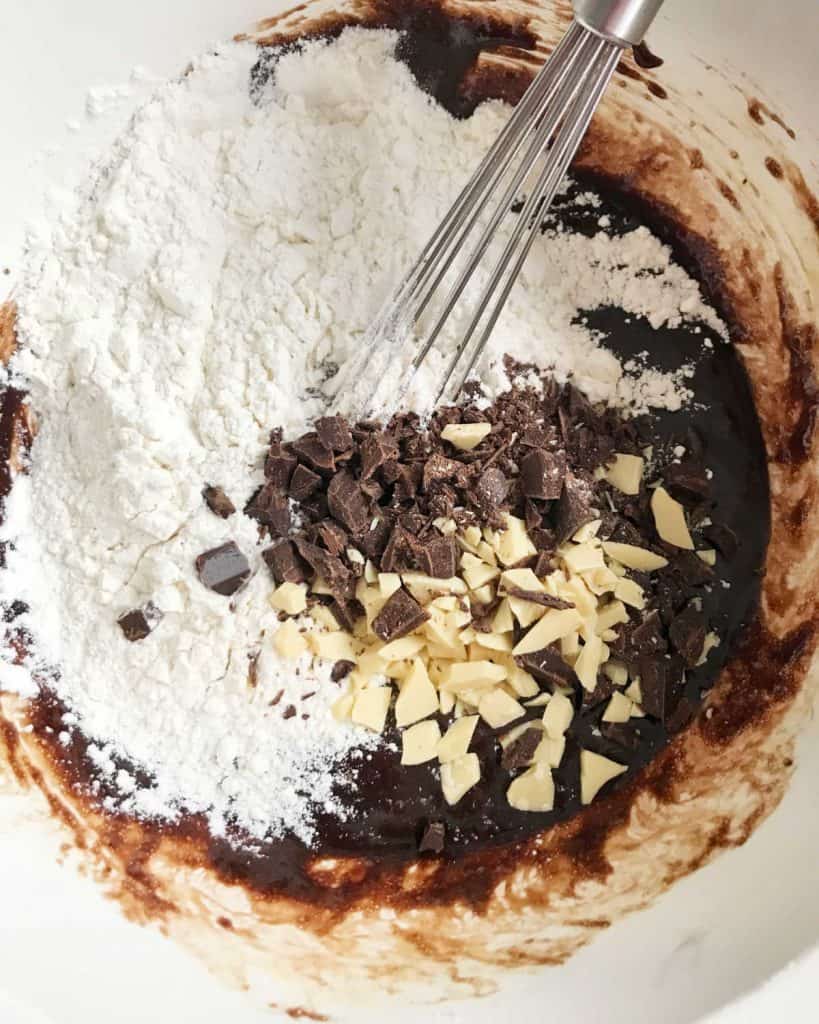 Flour, white and dark chocolate chunks in a bowl with a wire whisk. 