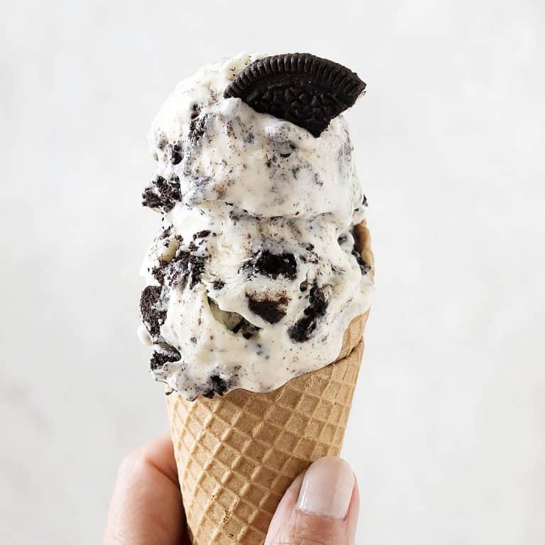Waffle cone with two scoops of Oreo ice cream being held on a light grey background.