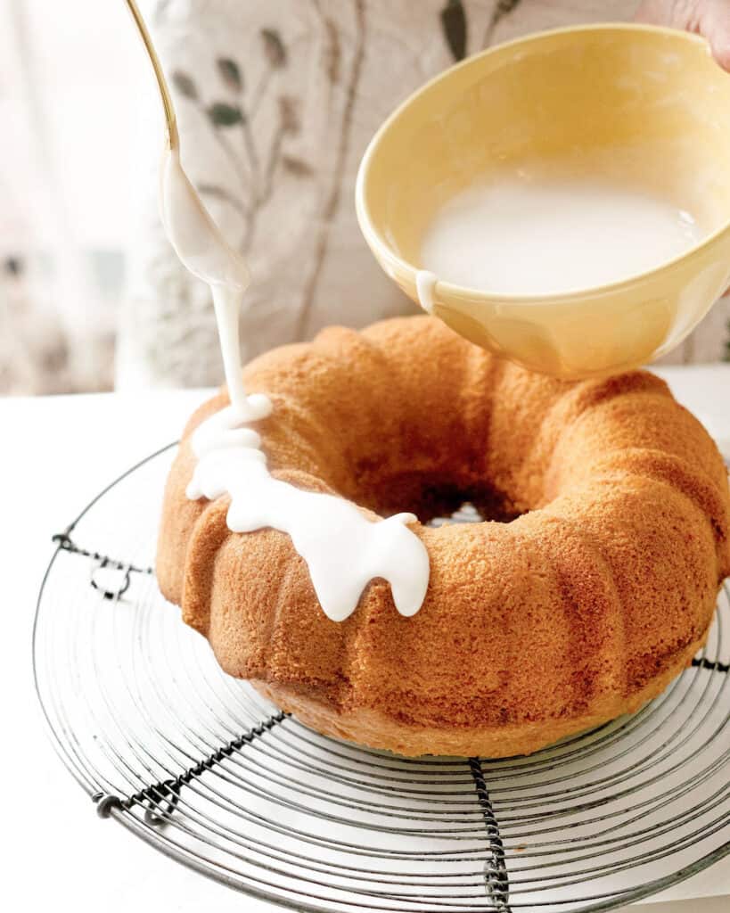 Drizzling lemon glaze from a yellow bowl onto a bundt cake on a wire rack.