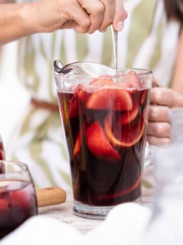Glass pitcher with sangria being stirred by person with green and white dress.