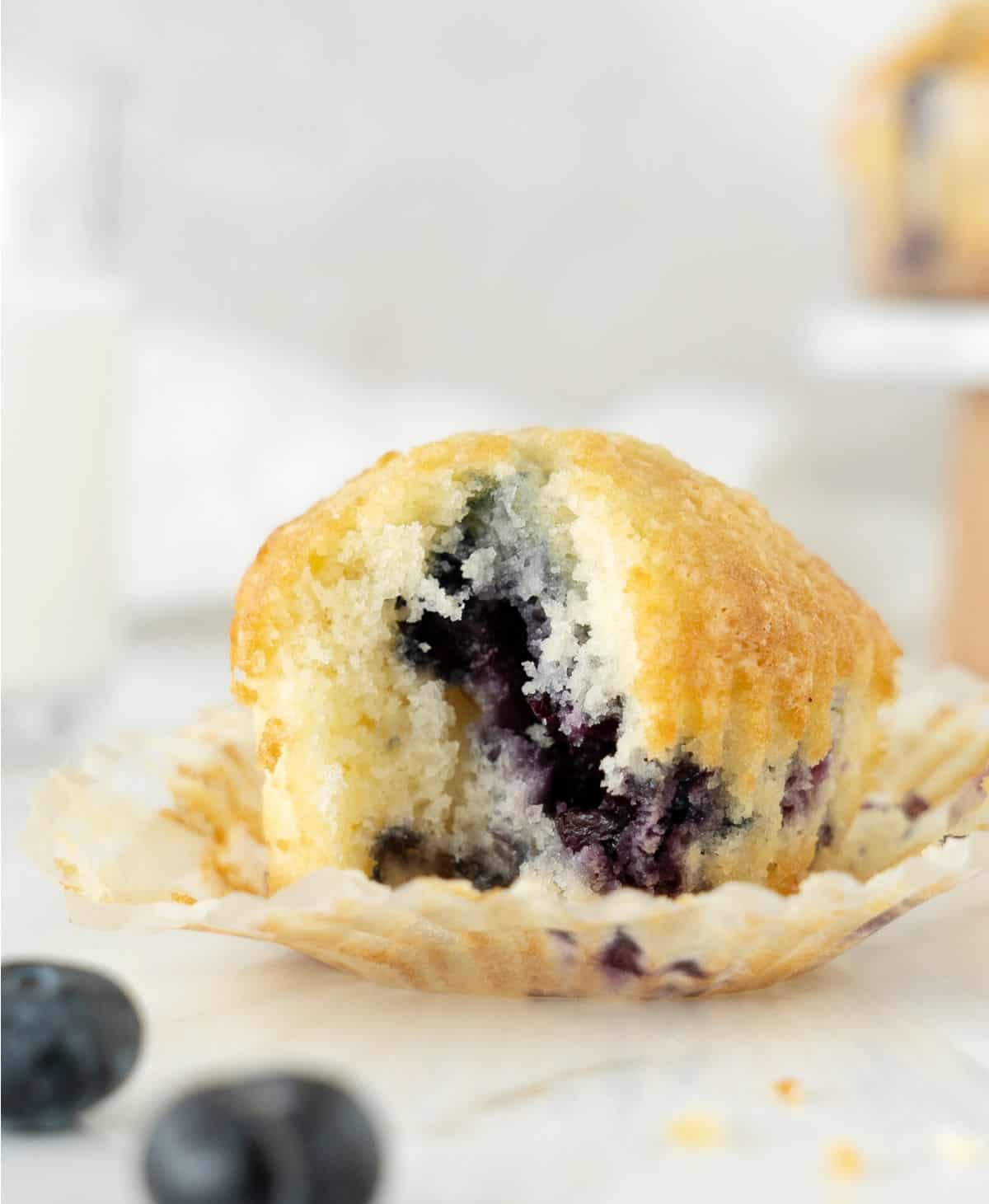 Bitten blueberry muffin in an opened paper liner. White marble surface, light grey background.