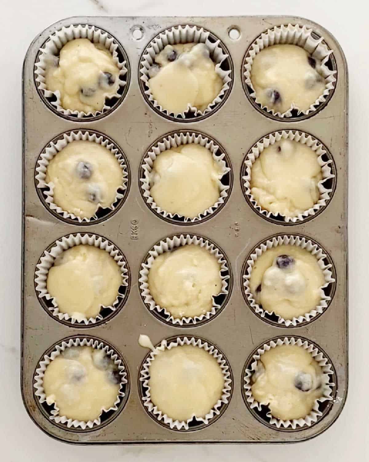 Muffin pan with blueberry muffin batter in paper liners. White marble surface.