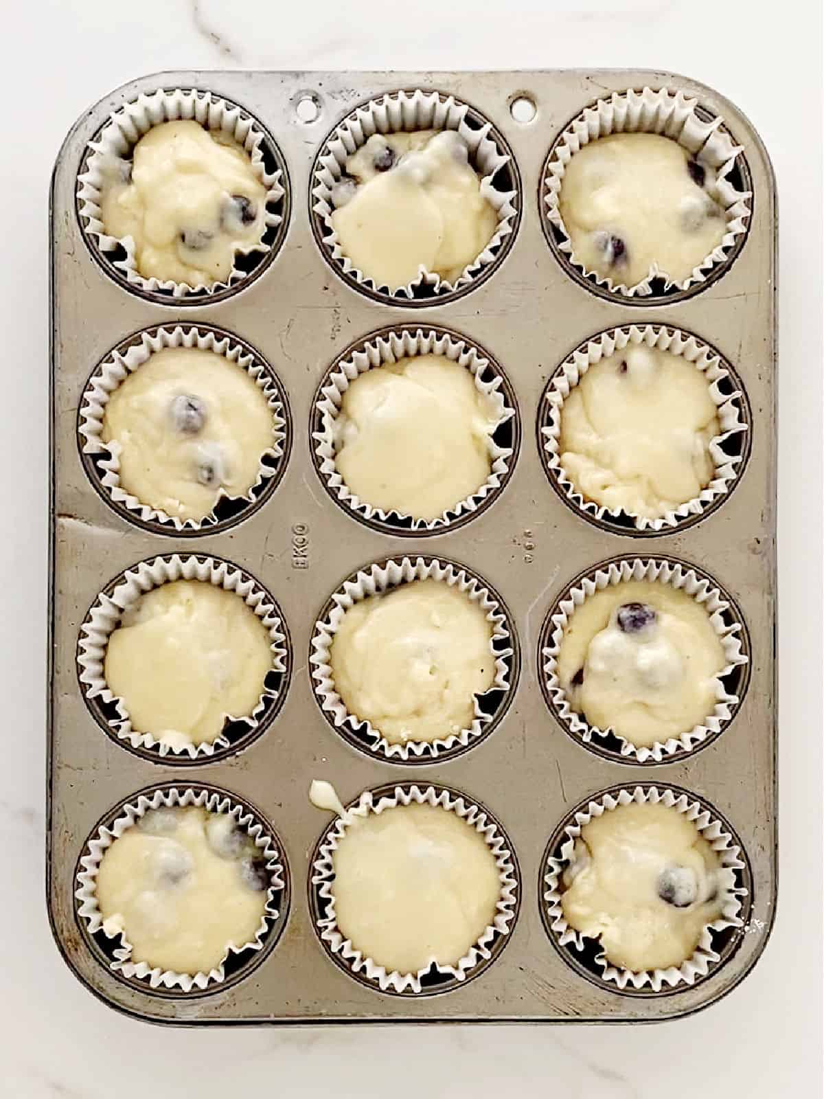 Muffin pan with blueberry muffin batter in paper liners. White marble surface.