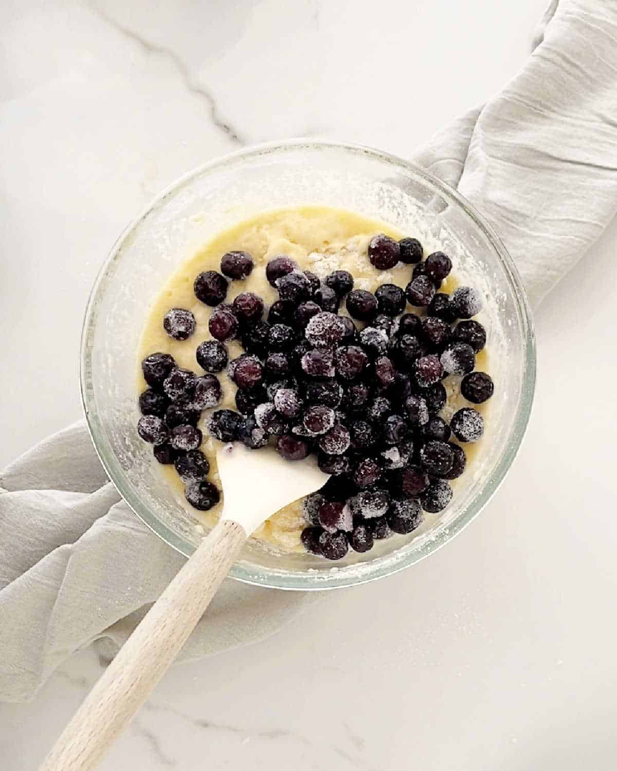 Blueberries added to muffin batter in a glass bowl with a spatula. White marble surface with light grey cloth. 