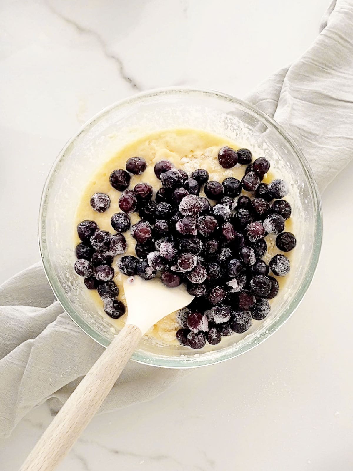 Blueberries added to muffin batter in a glass bowl with a spatula. White marble surface with light grey cloth.