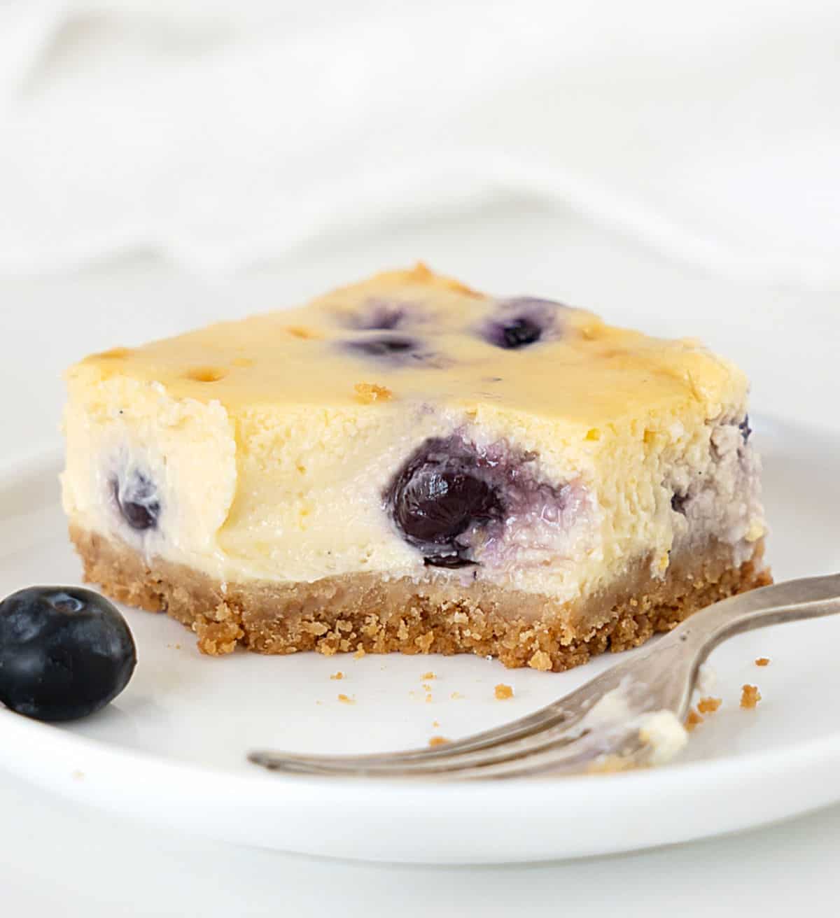 Eaten square of blueberry cheesecake bars on a white plate with a silver fork. White background. 