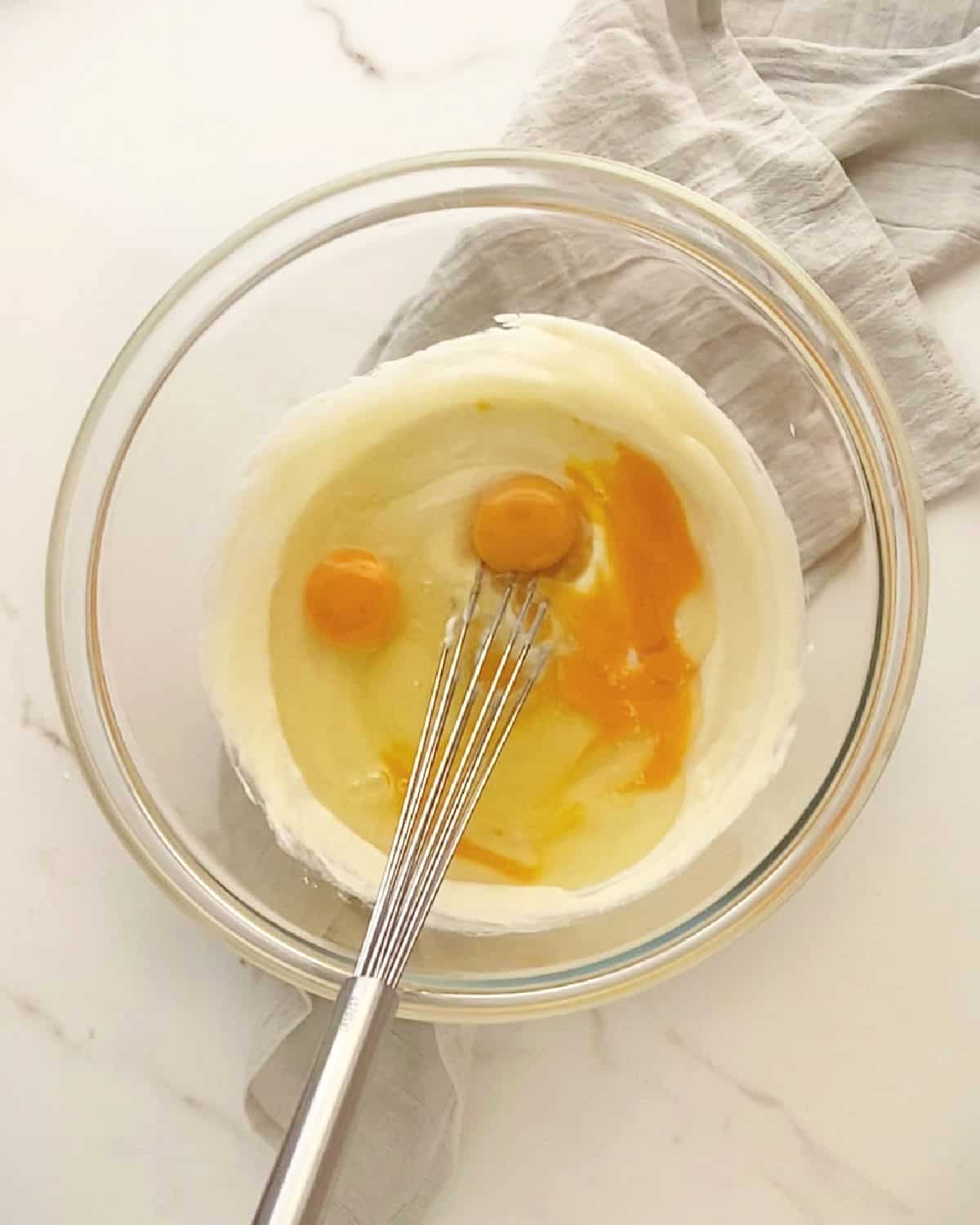 Glass bowl with whisk, cheesecake mixture and eggs on white marble with grey cloth. 
