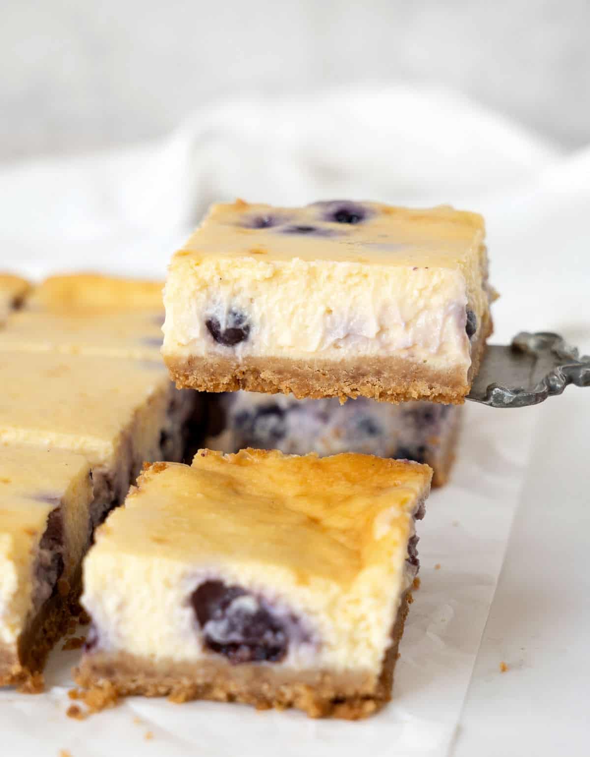 Squares of blueberry cheesecake on a white paper, one is being lifted. White background.