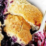 Purple and brown text overlay on serving of blueberry cobbler in a white bowl with a spoon.