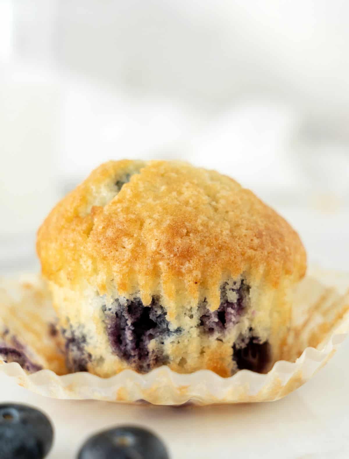 Single blueberry muffin in an opened paper liner. White surface, grey white background.