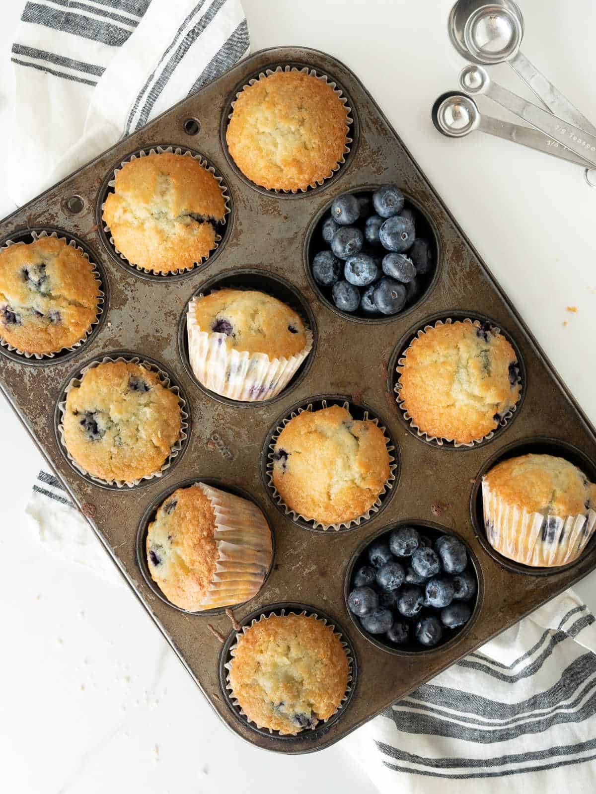 Several blueberry muffins in the metal pan with fresh blueberries. White surface, grey striped cloth. Top view. 