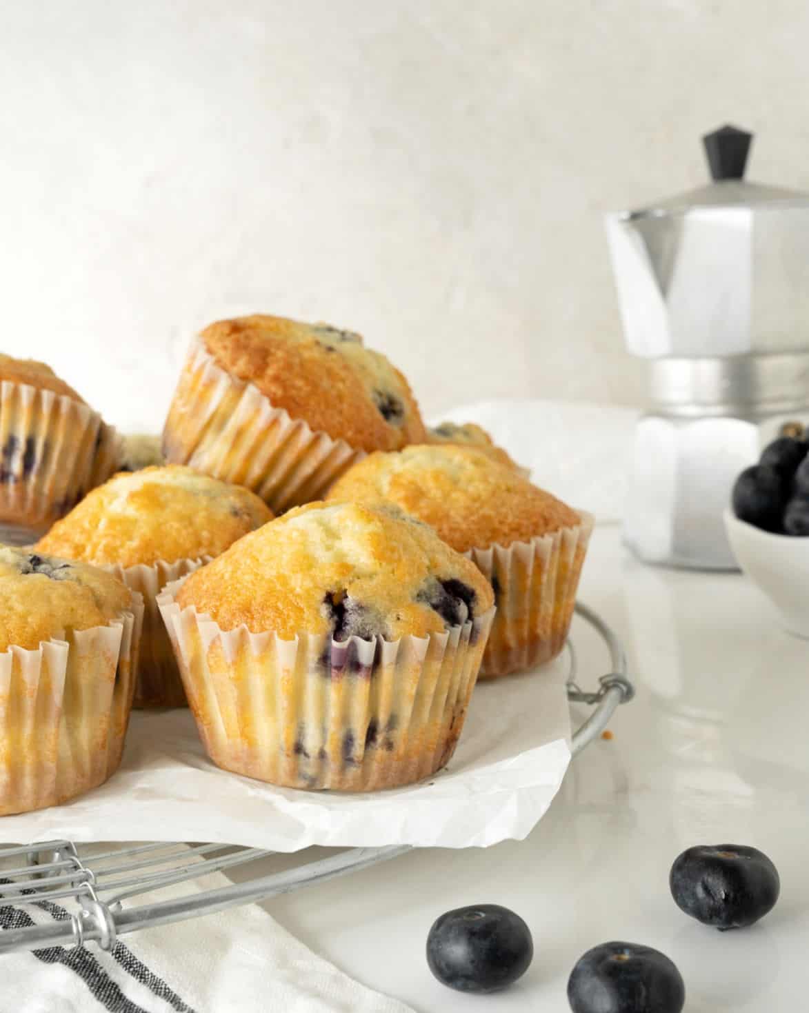 Grey background, white marble surface with pile of blueberry muffins on a wire rack with a white paper. Metal coffee pot, loose blueberries. 