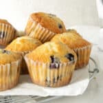 Several blueberry muffins in paper liners on a wire rack with parchment paper. Light grey background.