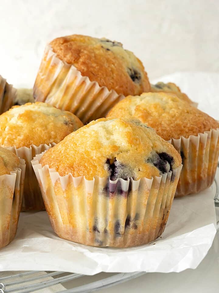 Several blueberry muffins in paper liners on a wire rack with parchment paper. Light grey background.