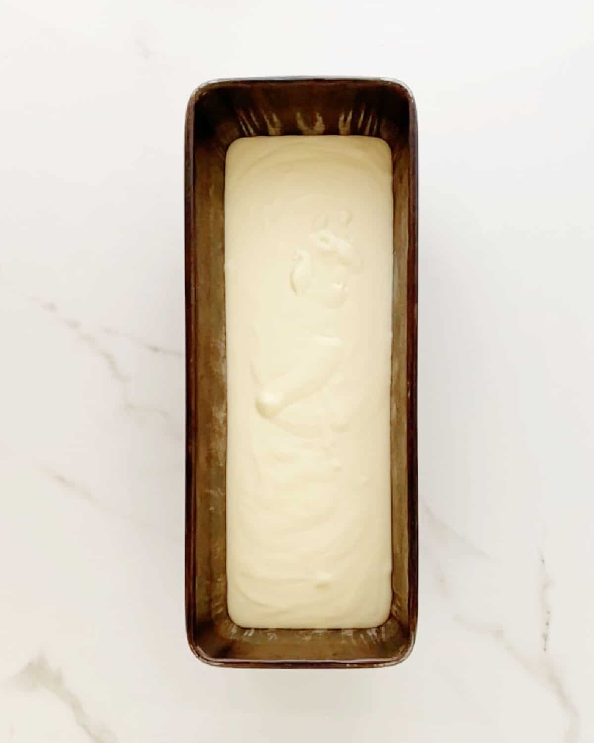 Dark loaf pan with ice crema mixture on a white marbled surface.